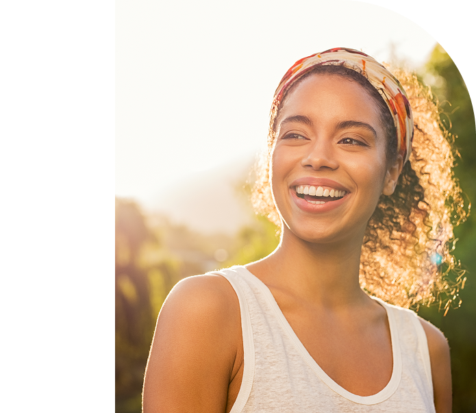Smiling person outdoors in sunlight, wearing a white tank top and headband, with a blurred background.